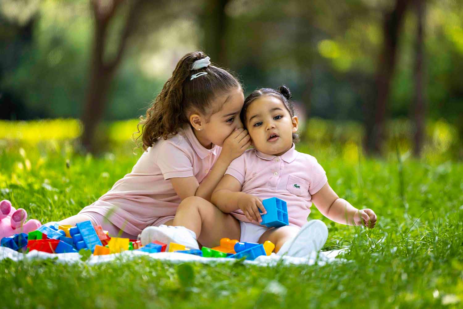 Deux petites filles en tenues roses jouent avec des blocs de construction sur l'herbe, capturées par un photographe lors d'une séance photo en extérieur.