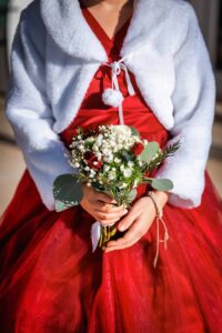 Demoiselle d'honneur en robe rouge et cape blanche tenant un bouquet de fleurs d'hiver.