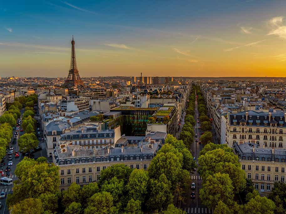 Drone 16 Vue aérienne de Paris au crépuscule, avec la Tour Eiffel et les avenues bordées d'arbres en évidence, sous un ciel aux teintes orange et bleu.