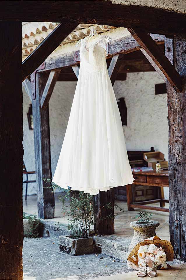 Robe de mariée suspendue avant les préparatifs Robe de mariée suspendue sous une poutre en bois avec bouquet et chaussures au sol