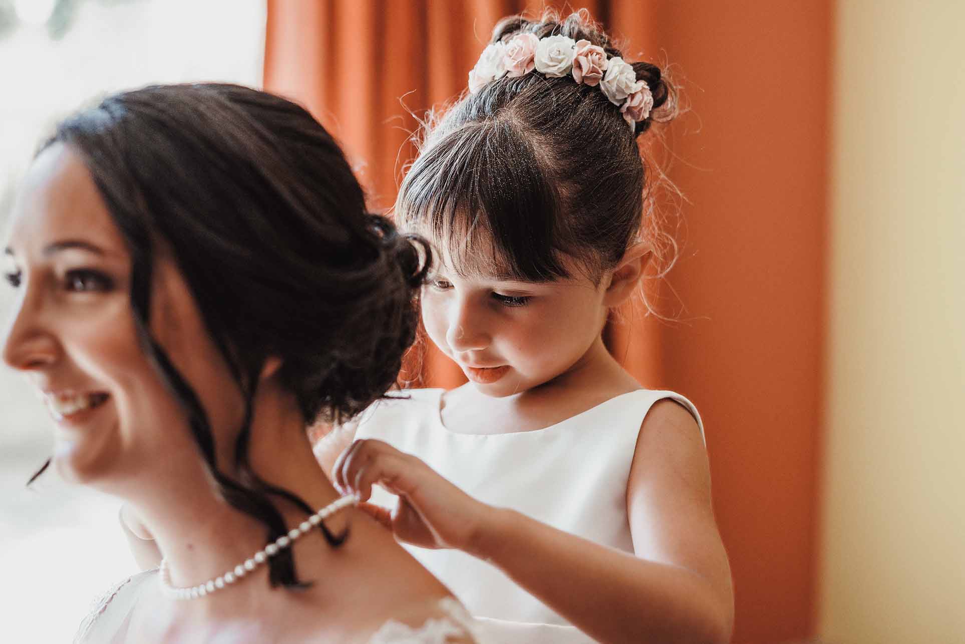 Un moment complice pendant les préparatifs de la mariée Une enfant aide la mariée à mettre son collier pendant les préparatifs